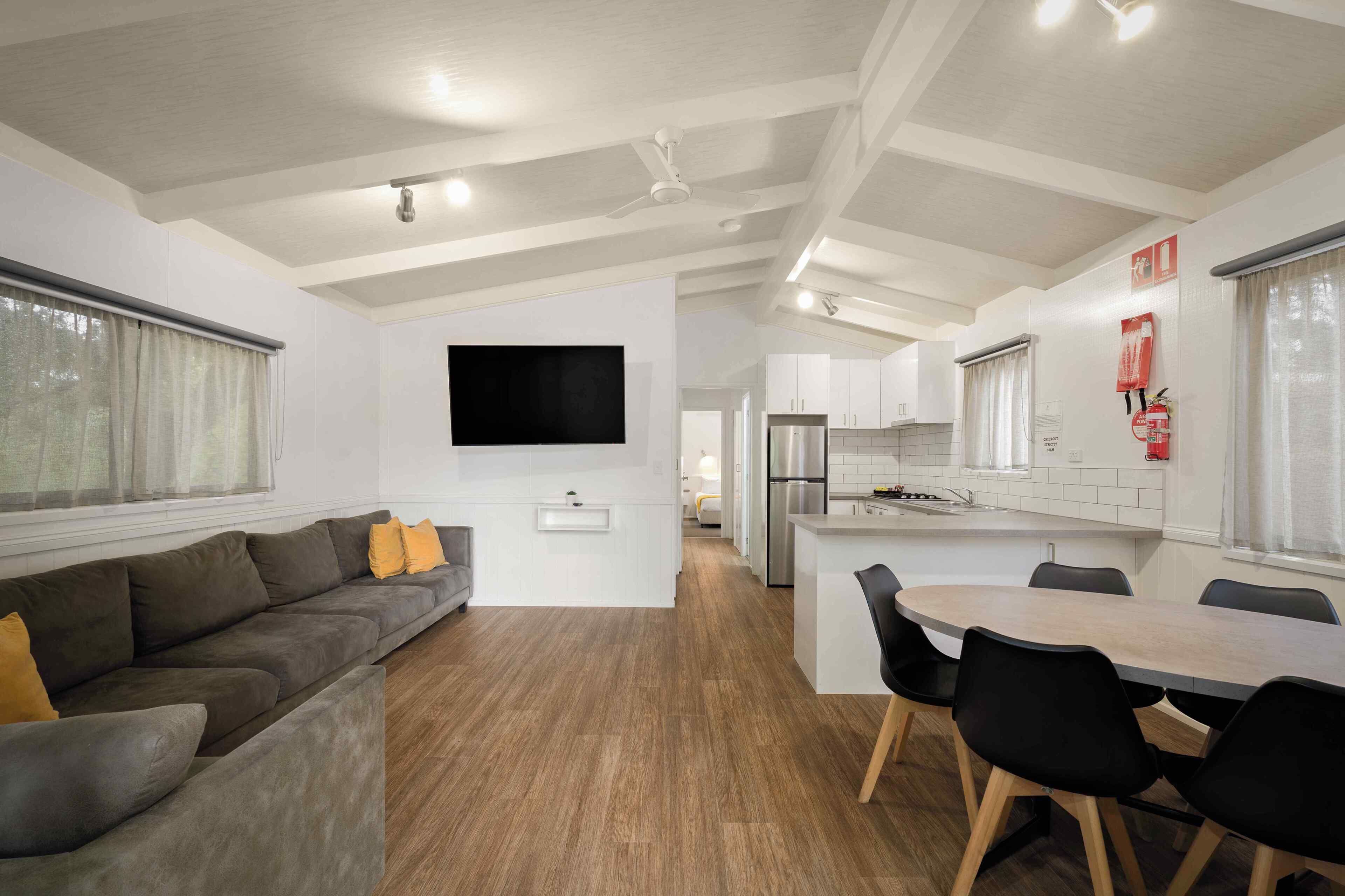 A modern living room with a sofa, a dining table with chairs, a kitchen area, and a flat-screen TV mounted on the wall.