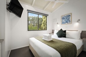 Habitación de hotel con cama, ventana con vista a árboles, ventilador de techo y televisión montada en la pared.