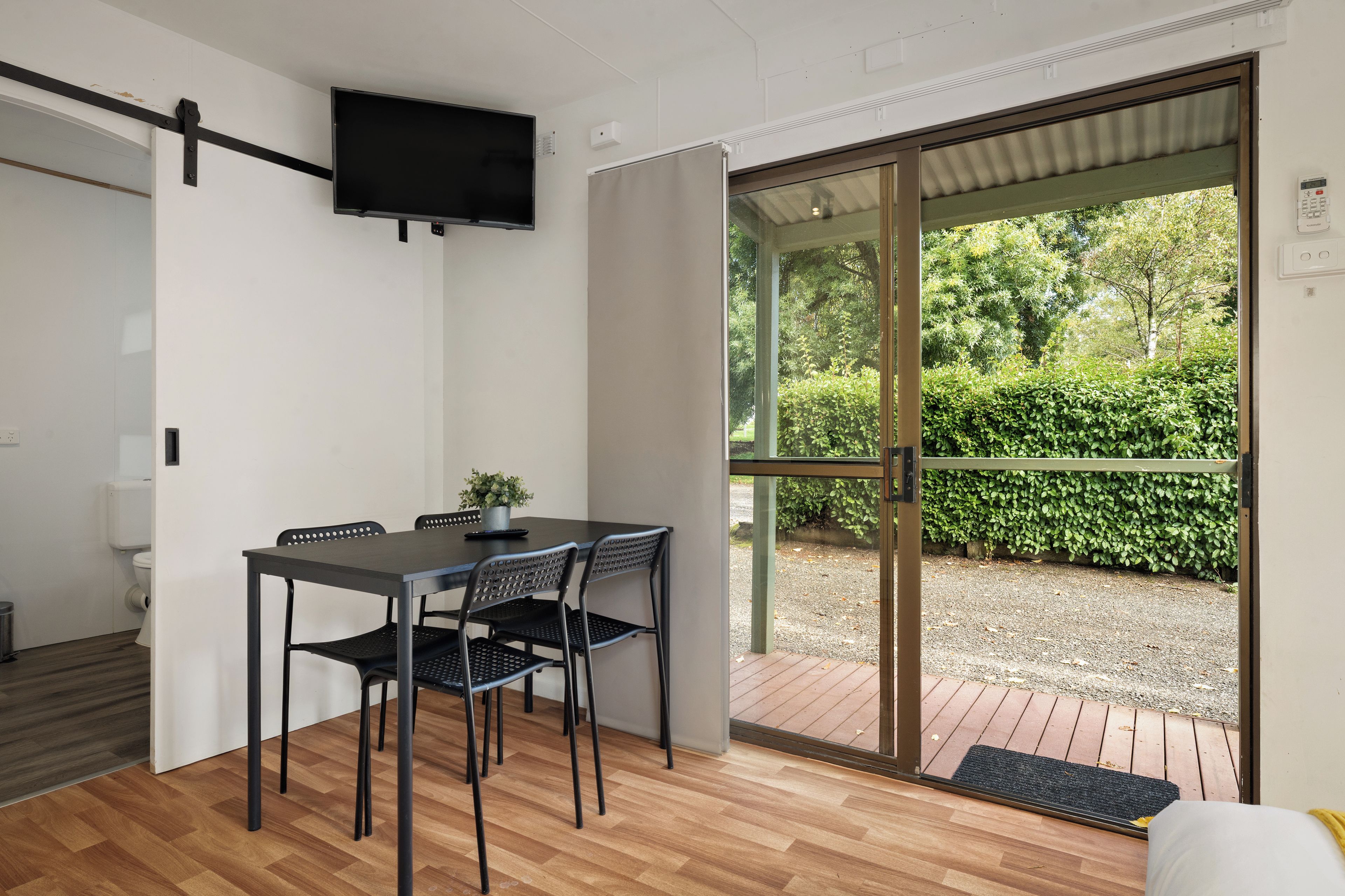 A modern room with a dining table, chairs, a television, and a sliding glass door leading to an outdoor area with greenery.