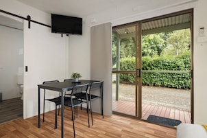 A modern room with a dining table, chairs, a television, and a sliding glass door leading to an outdoor area with greenery.