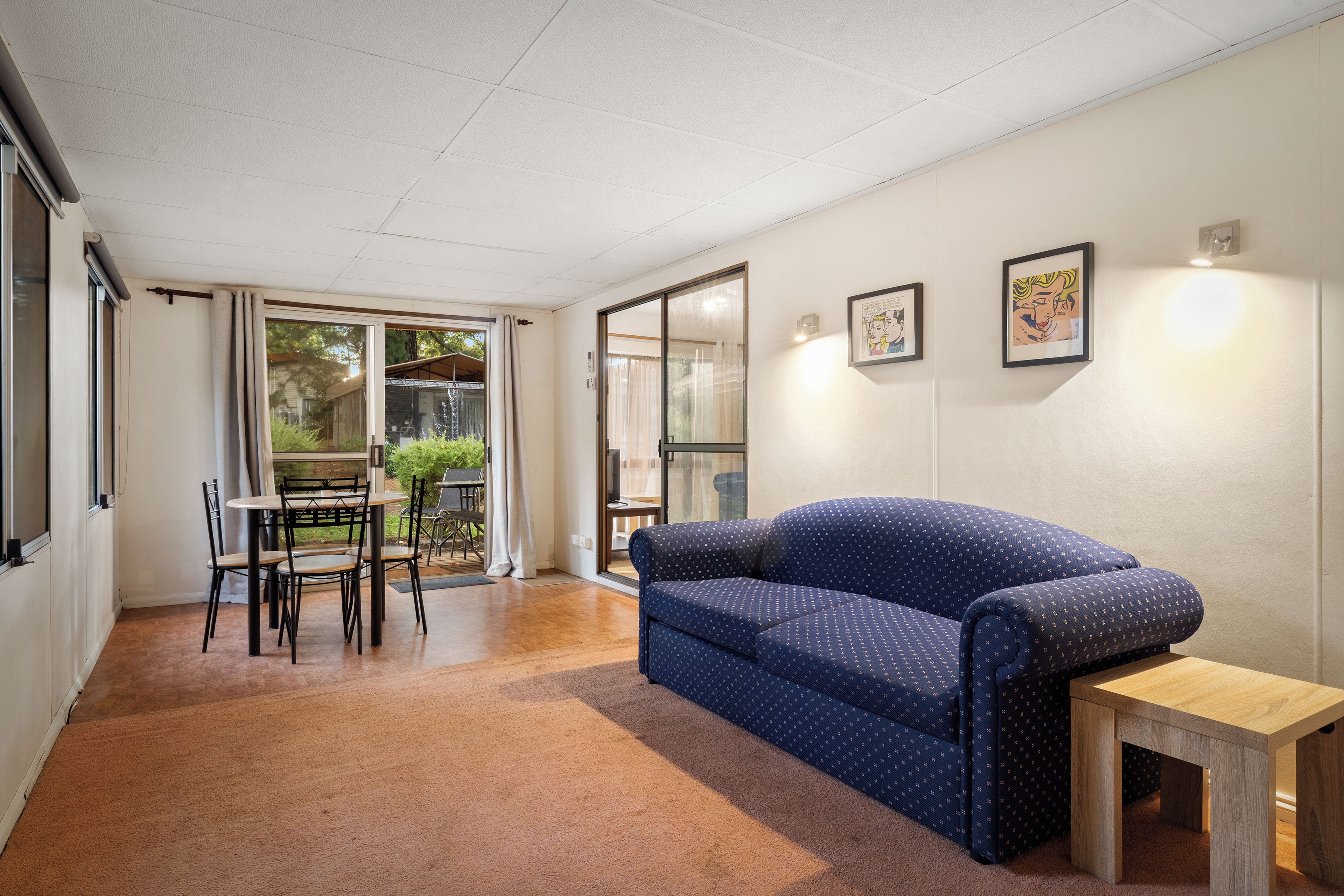 Una habitación con un sofá azul, una pequeña mesa de madera, una mesa de comedor con sillas y un amplio ventanal con cortinas.