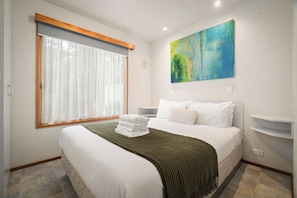 Une chambre d’hôtel avec un lit, une fenêtre avec des rideaux, un tableau au mur et une table de chevet.