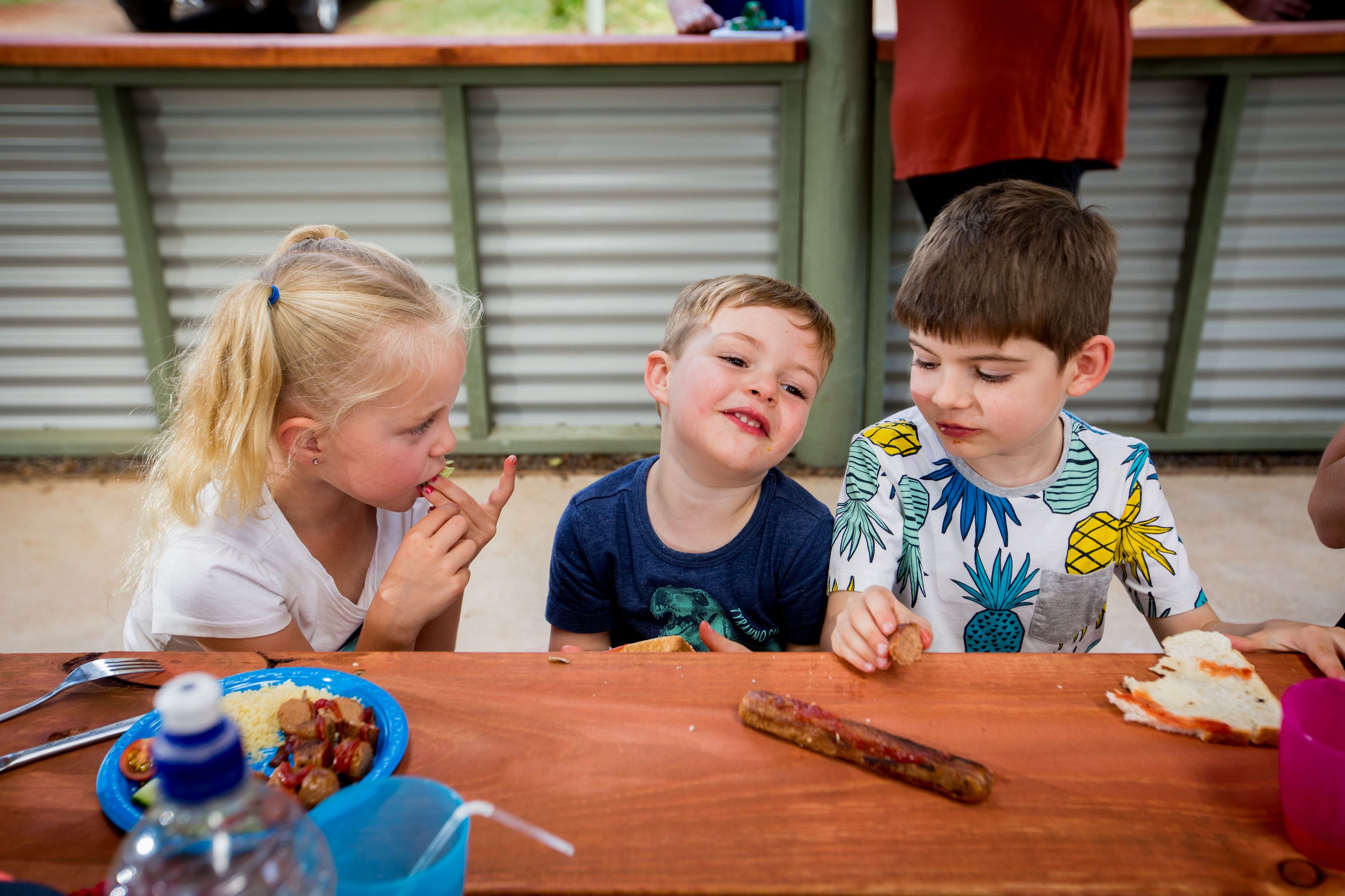 A group of children sitting at a wooden table with food items and a water bottle.