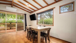 Un coin repas avec une table et des chaises en bois, un téléviseur fixé au mur et de grandes fenêtres donnant sur un paysage verdoyant.