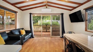 Une pièce avec un plafond en bois, une porte-fenêtre coulissante donnant sur une terrasse extérieure, un canapé en cuir noir, une table à manger en bois et un téléviseur à écran plat fixé au mur.