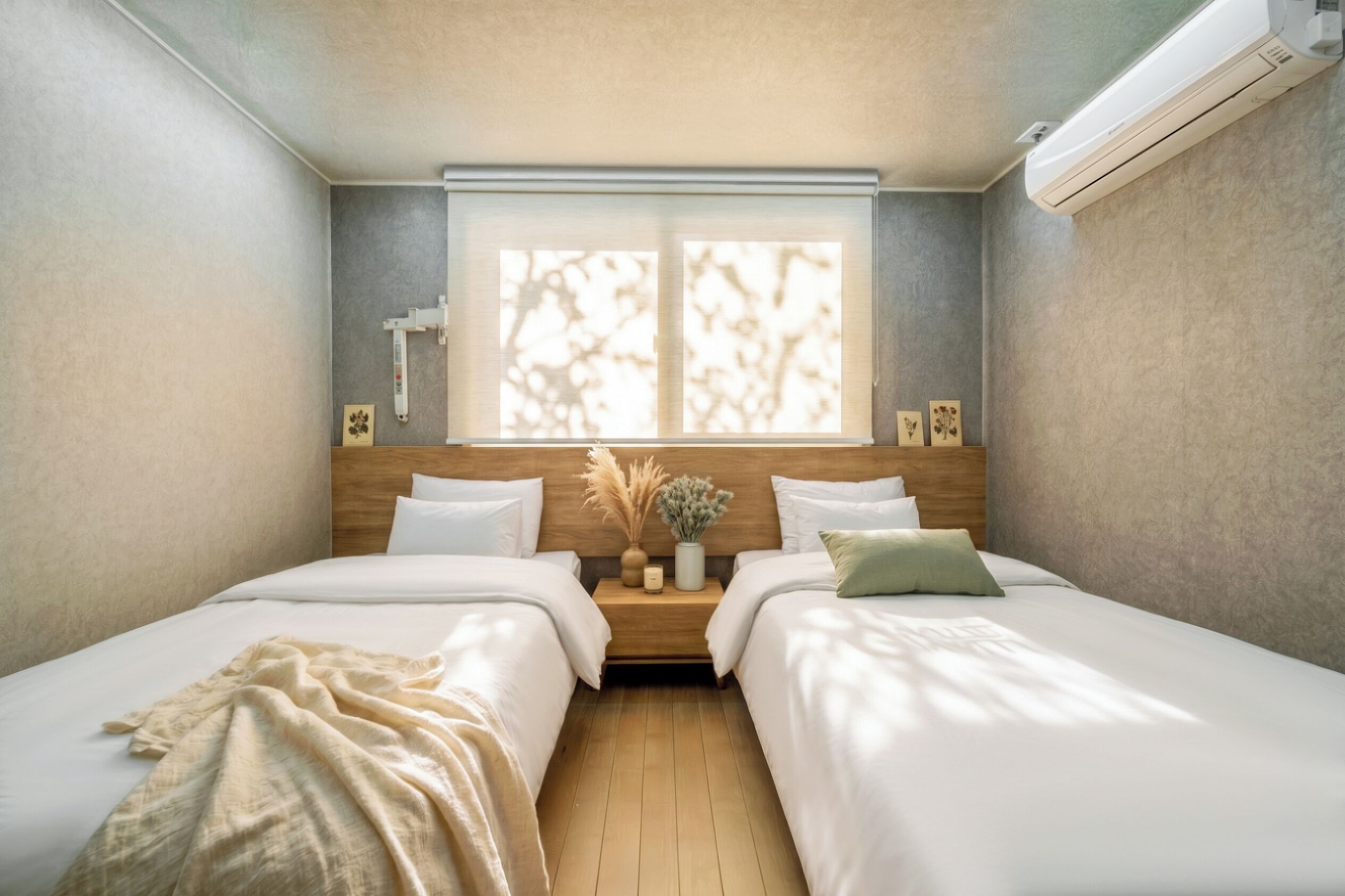 A hotel room with two beds, a window with blinds, a nightstand with a vase and candles, and an air conditioning unit.