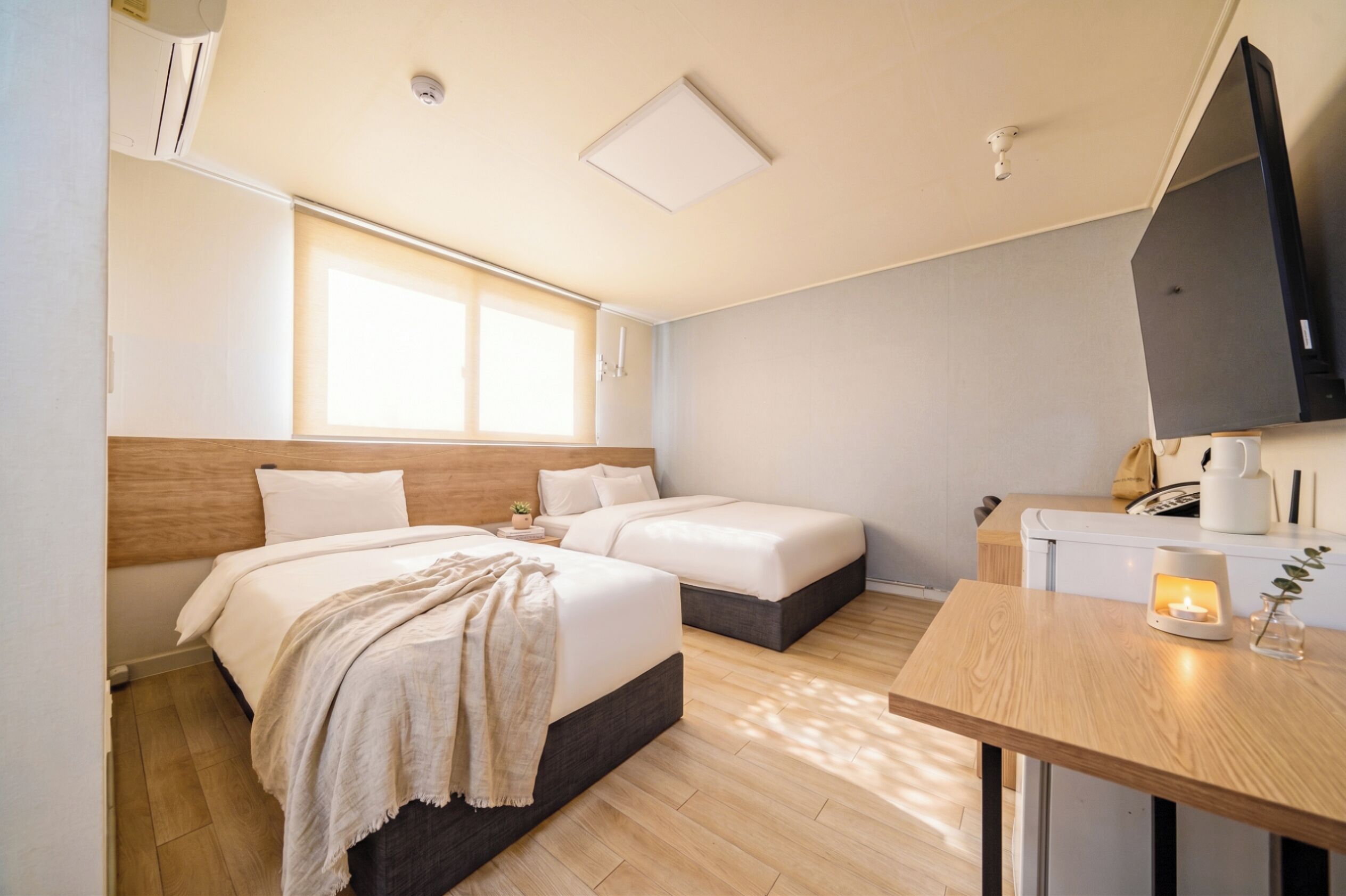 A hotel room with two beds, a wooden headboard, a bedside table with a candle, a TV, and a window with blinds.