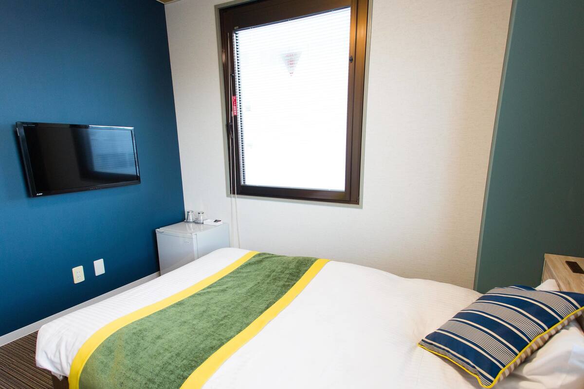 A hotel room with a bed, a TV mounted on the wall, a window, and a small bedside table.