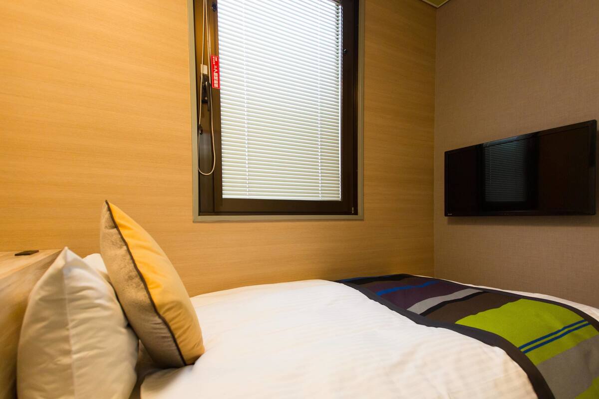 A hotel room with a bed, pillows, a window with blinds, and a flat-screen TV mounted on the wall.