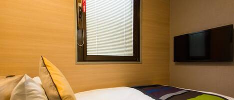 Ein Hotelzimmer mit Bett, Kissen, einem Fenster mit Jalousien und einem an der Wand befestigten Flachbildfernseher.
