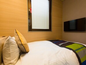 A hotel room with a bed, pillows, a window with blinds, and a flat-screen TV mounted on the wall.