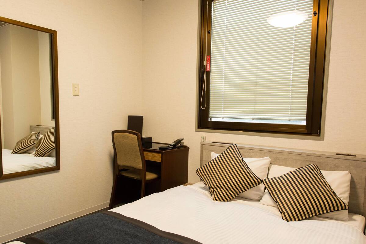 A hotel room with a bed, a desk, a chair, a mirror, and a window with blinds.