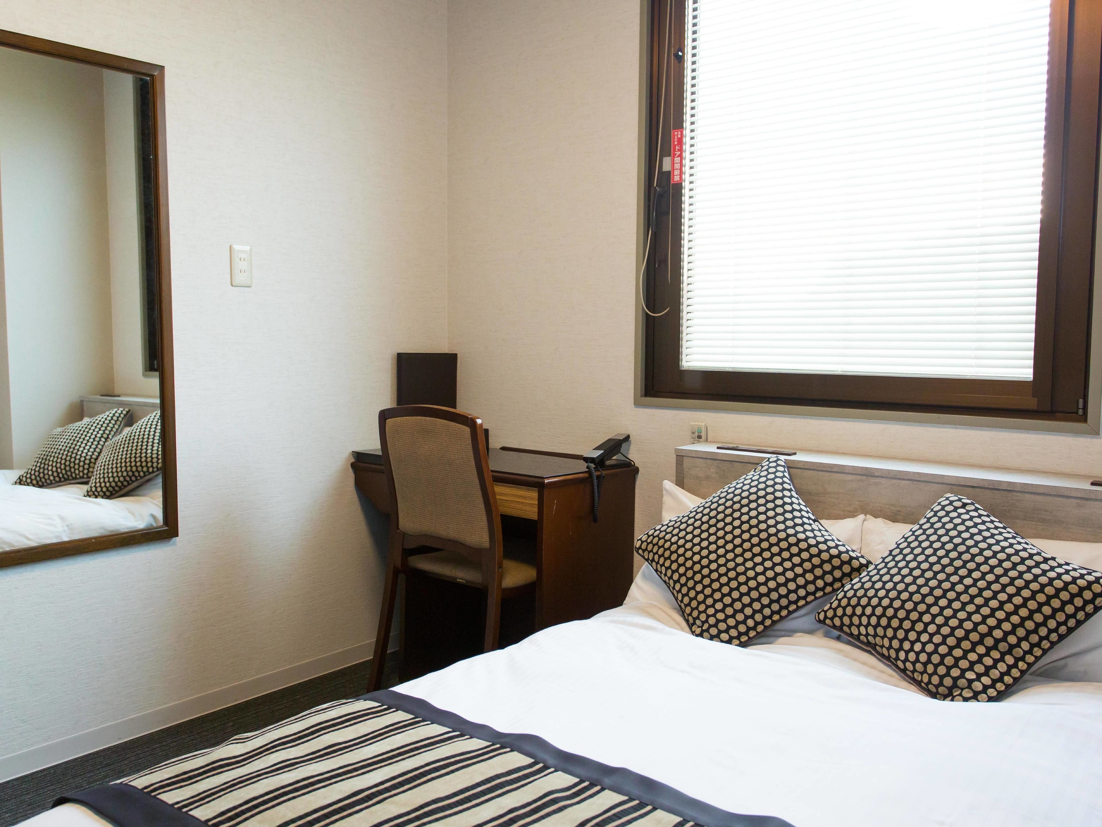 A hotel room with a bed, a desk, a chair, a mirror, and a window with blinds.