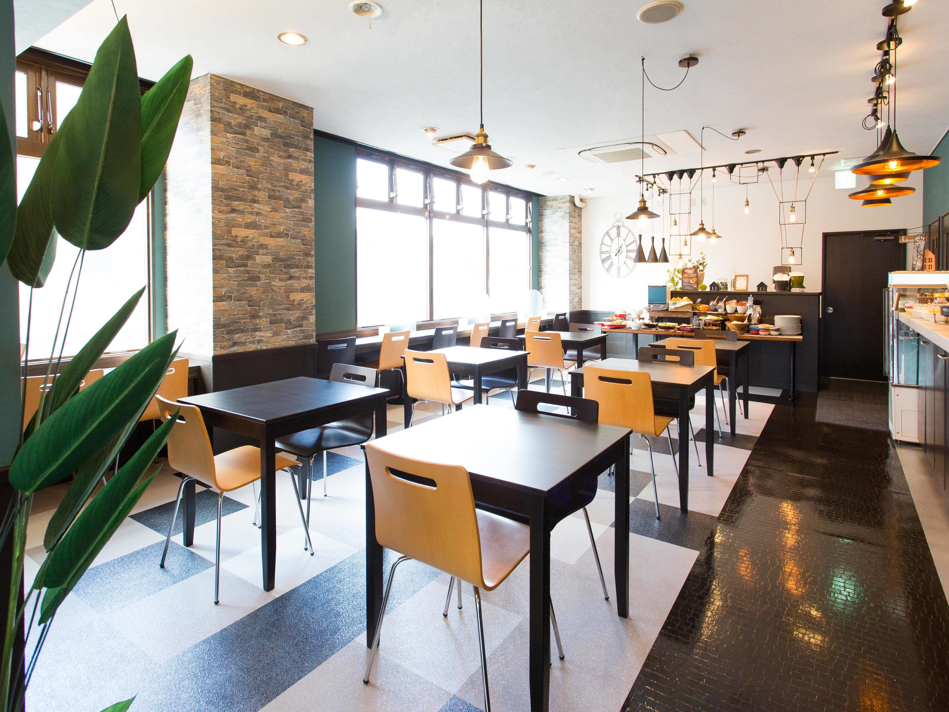 A modern dining area with tables and chairs, a kitchen in the background, and large windows allowing natural light.