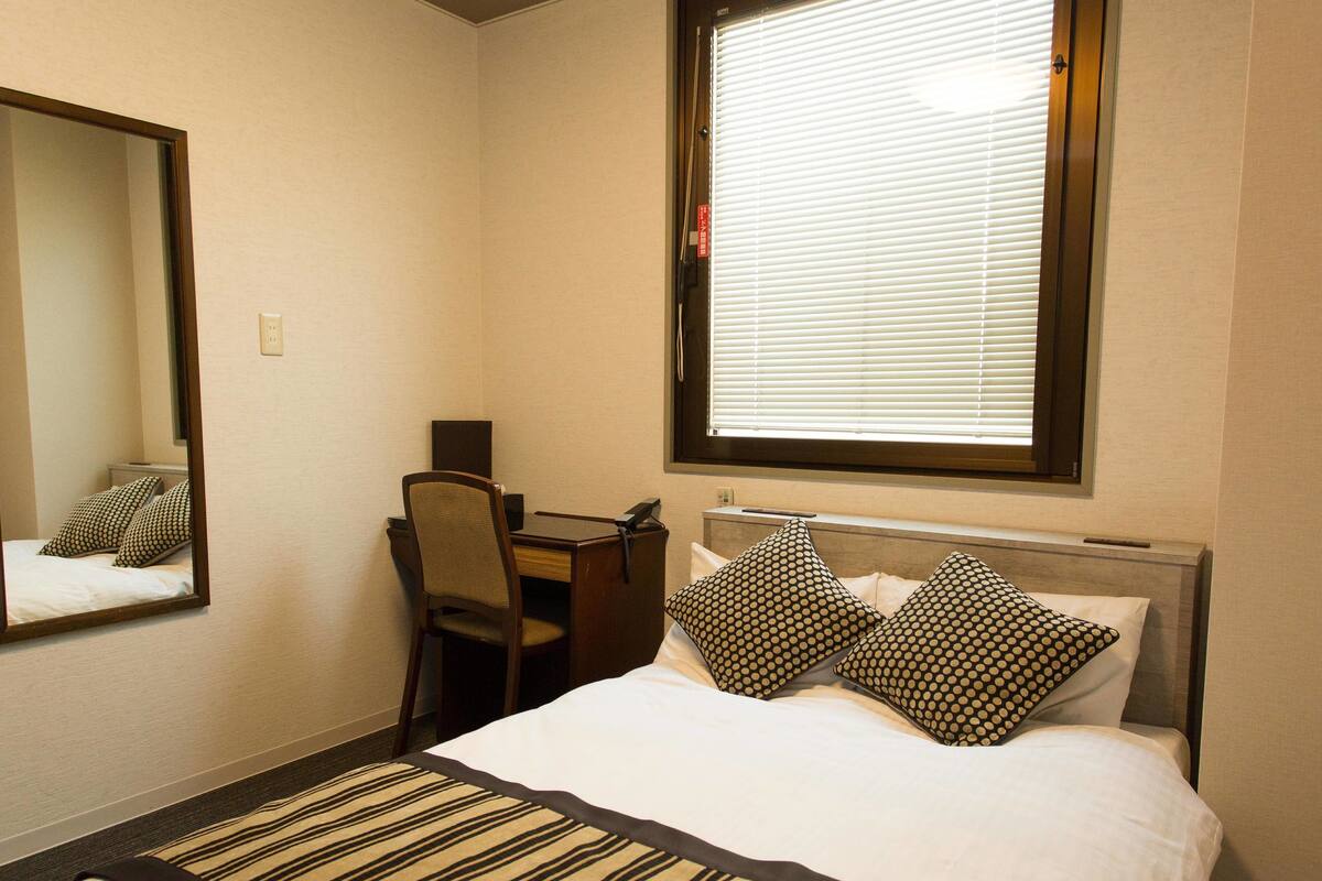 A hotel room with a bed, a desk, a chair, a mirror, and a window with blinds.