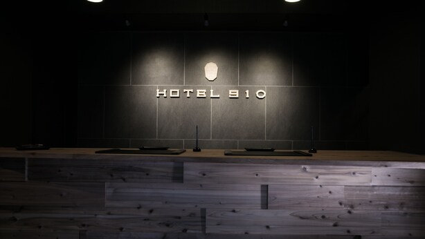 Eine moderne Hotelrezeption mit minimalistischem Design, ein dunkler Wandbereich mit der Hotelbezeichnung in der Mitte beleuchtet.
