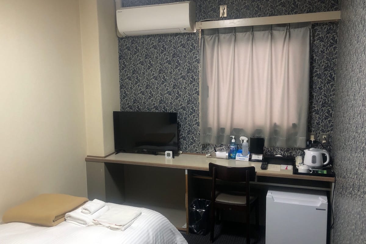 Habitación de hotel con cama, escritorio con televisor, silla, refrigerador pequeño y ventana con cortinas.