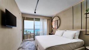 Habitación de hotel con una cama grande, un televisor de pantalla plana montado en la pared, un balcón con vistas al mar y un espejo redondo en la pared.