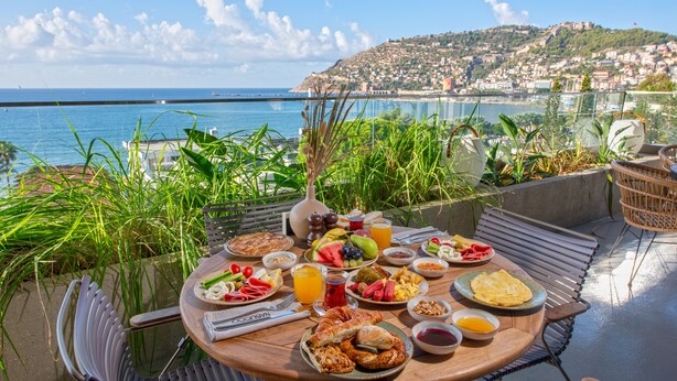 Una mesa redonda de madera con alimentos para el desayuno y vistas al mar y a una colina.