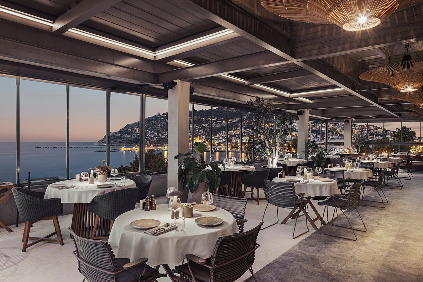 A restaurant with round tables, white tablecloths, and a view of a coastal city at dusk.