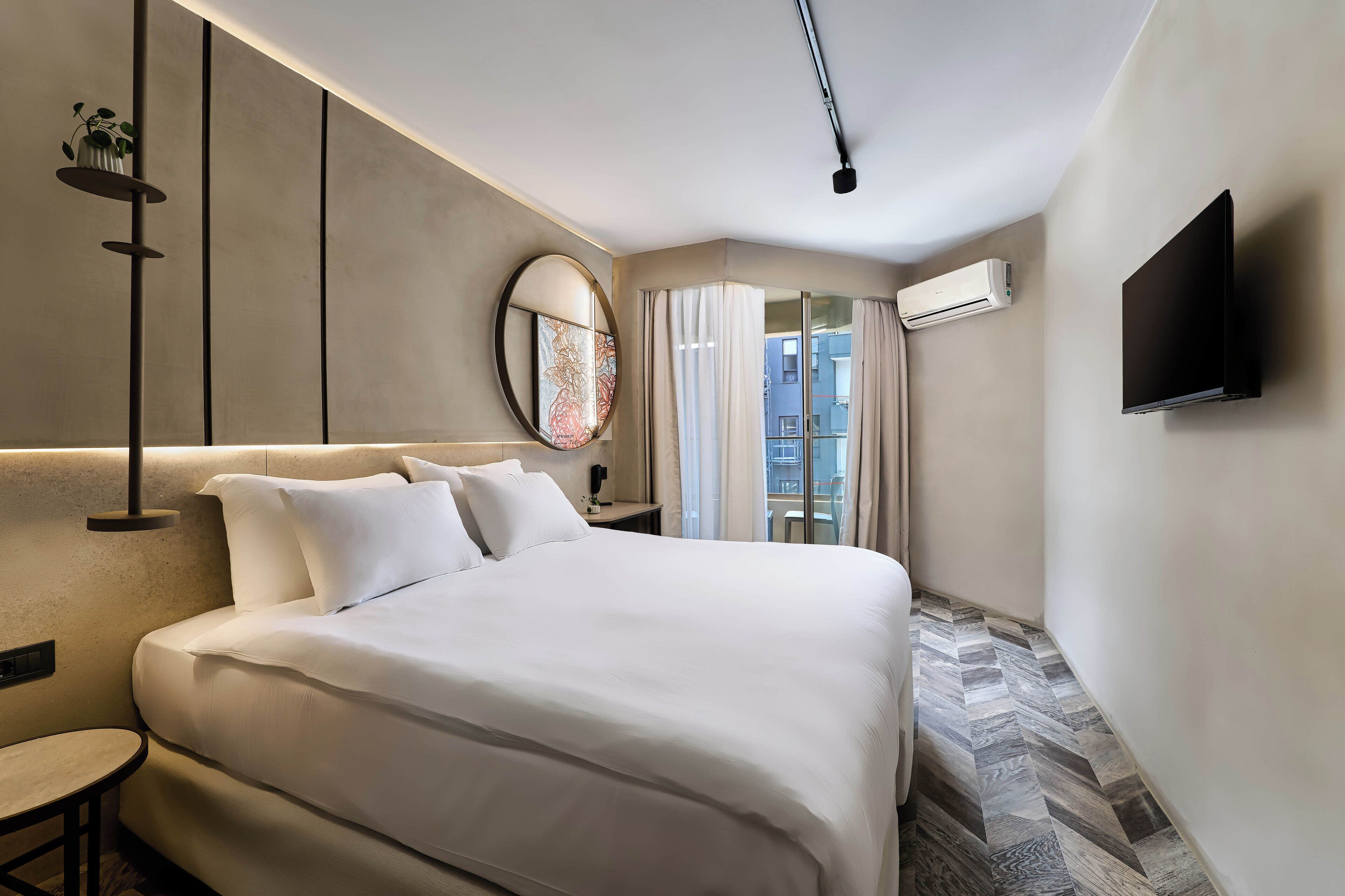A modern hotel room with a large bed, a bedside table, a round mirror, a flat-screen TV, and an air conditioning unit.