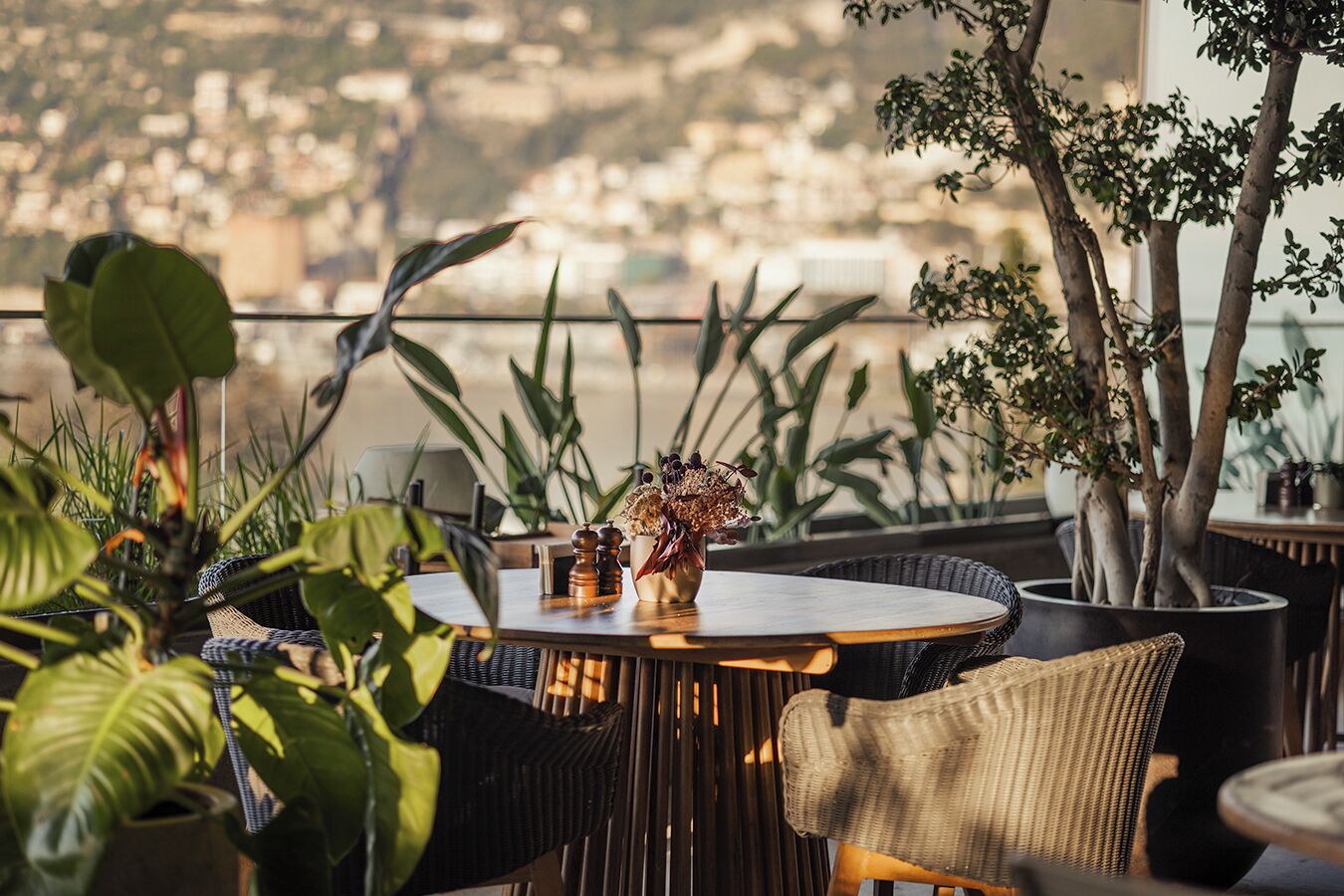 A round table with a vase of dried flowers, surrounded by wicker chairs, with a view of a cityscape in the background.