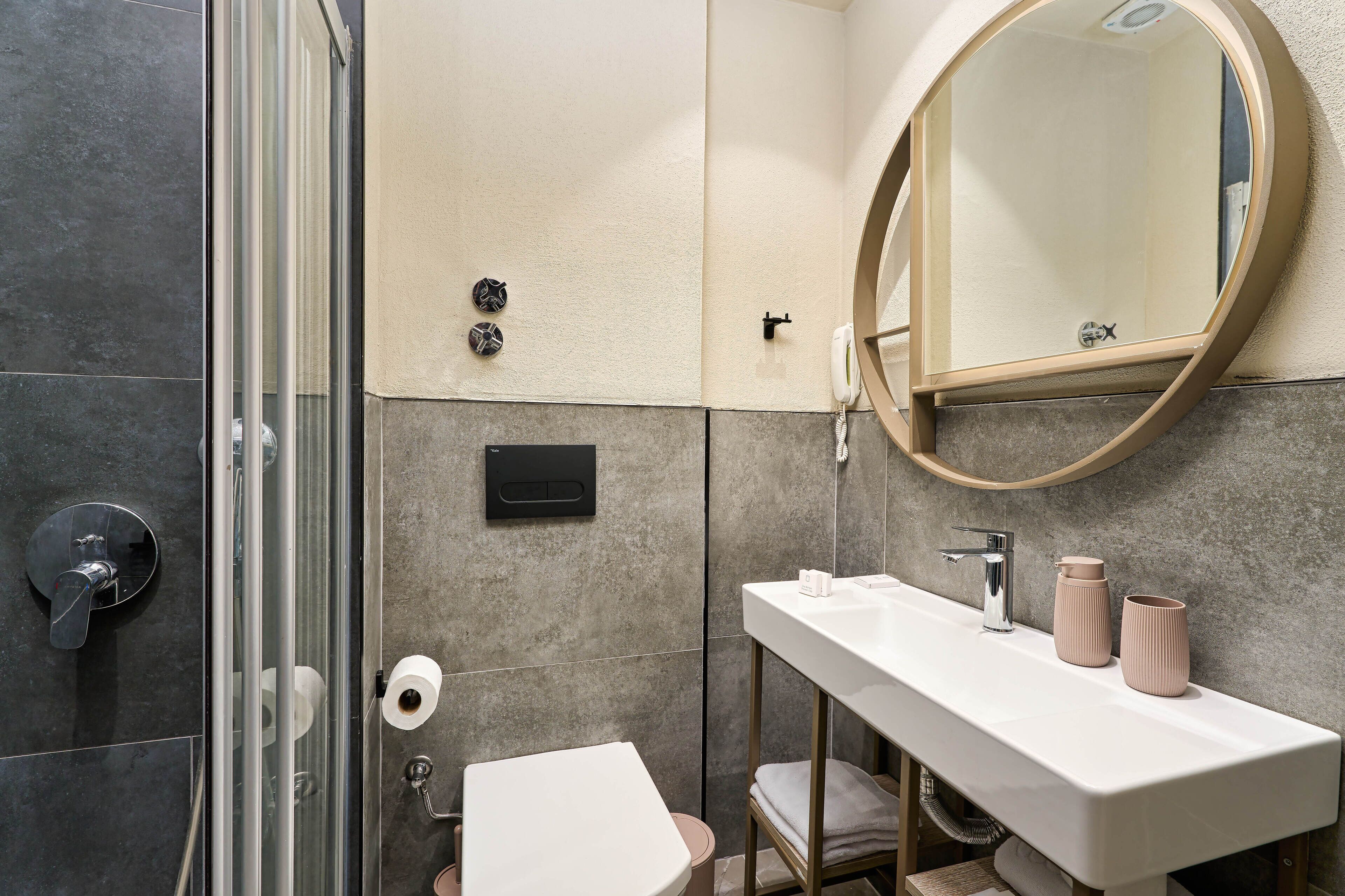 A bathroom with a glass shower enclosure, a white sink with two pink containers, and a round mirror.
