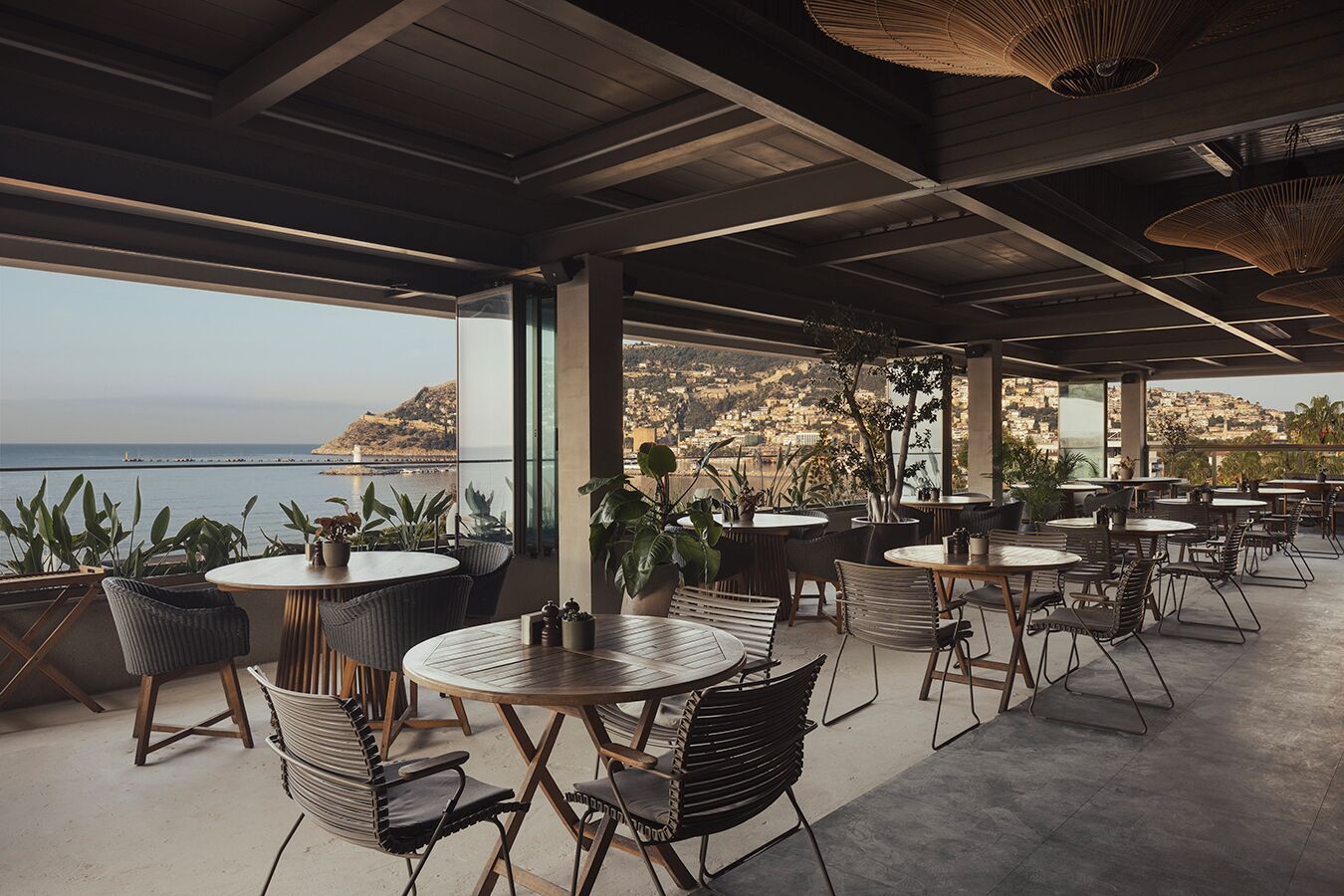 Un café en plein air avec des tables et des chaises rondes, de grandes fenêtres donnant sur la mer et sur une ville située sur une colline.