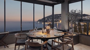 Comedor con una mesa dispuesta para cuatro personas, ventanas amplias con vistas a un pueblo costero y al mar.