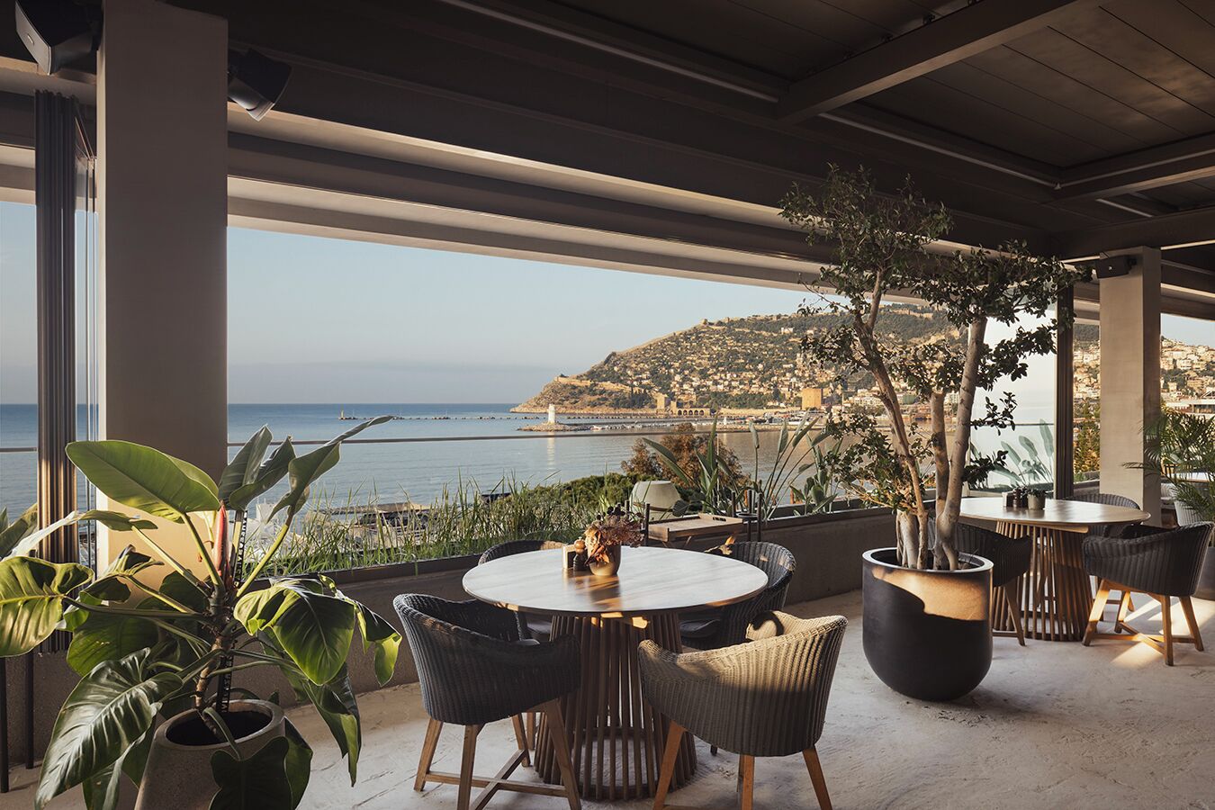 An outdoor seating area with a table and chairs, potted plants, and a view of the sea and a hillside town.