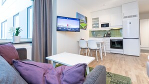 Een moderne woonkamer met een grijze bank, een witte tafel en een aan de muur bevestigde televisie. In de achtergrond is een keuken te zien met witte kasten, een magnetron en een koelkast.