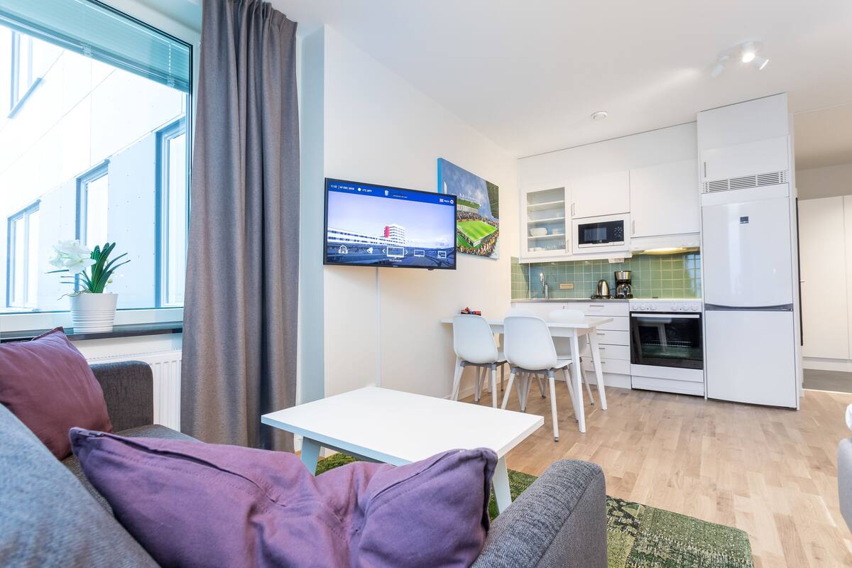 Una sala moderna con un sofá gris, una mesa blanca y un televisor montado en la pared. Se ve un área de cocina con armarios blancos, un microondas y un refrigerador al fondo.