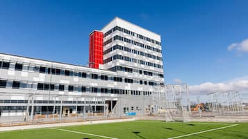 Un edificio moderno con fachada roja y blanca, una entrada de vidrio amplia y un campo verde frente a él.
