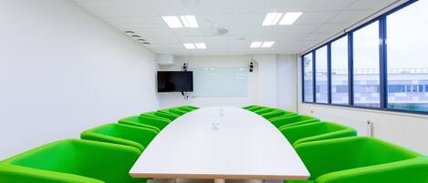 A conference room with a long white table and bright green chairs, large windows, and a flat-screen TV on the wall.