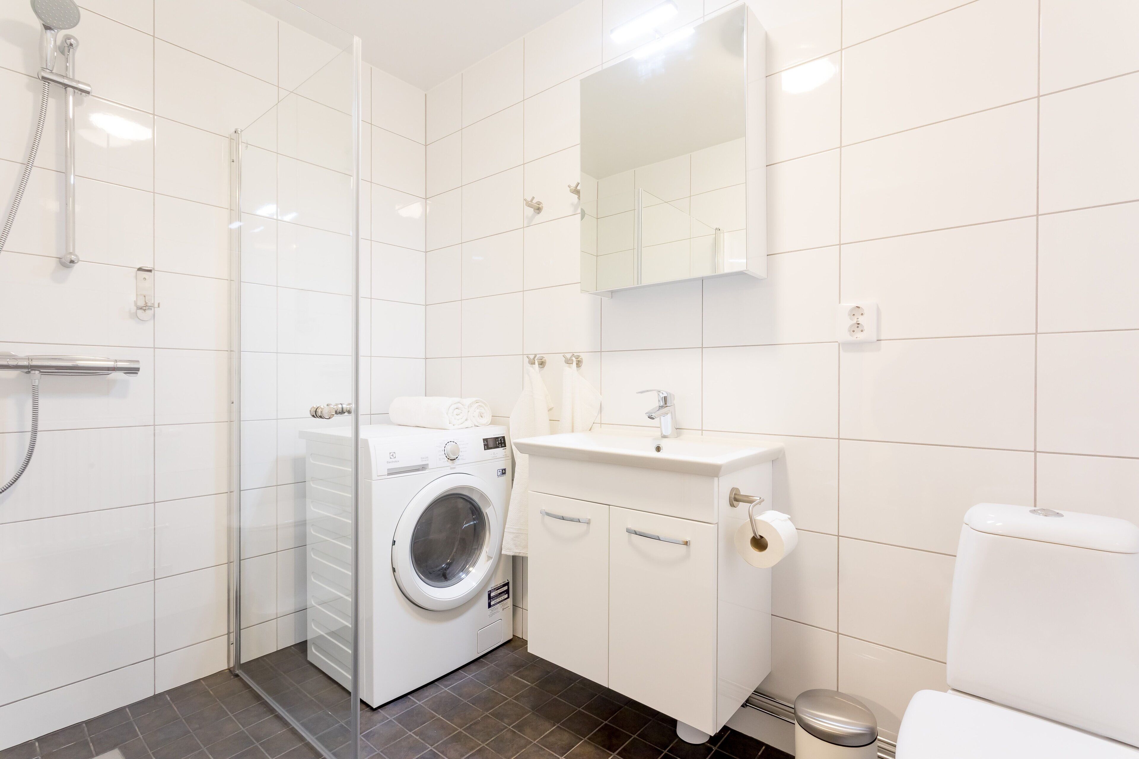 A modern bathroom with a glass-enclosed shower, a built-in washing machine, a sink with a mirror, and a toilet.