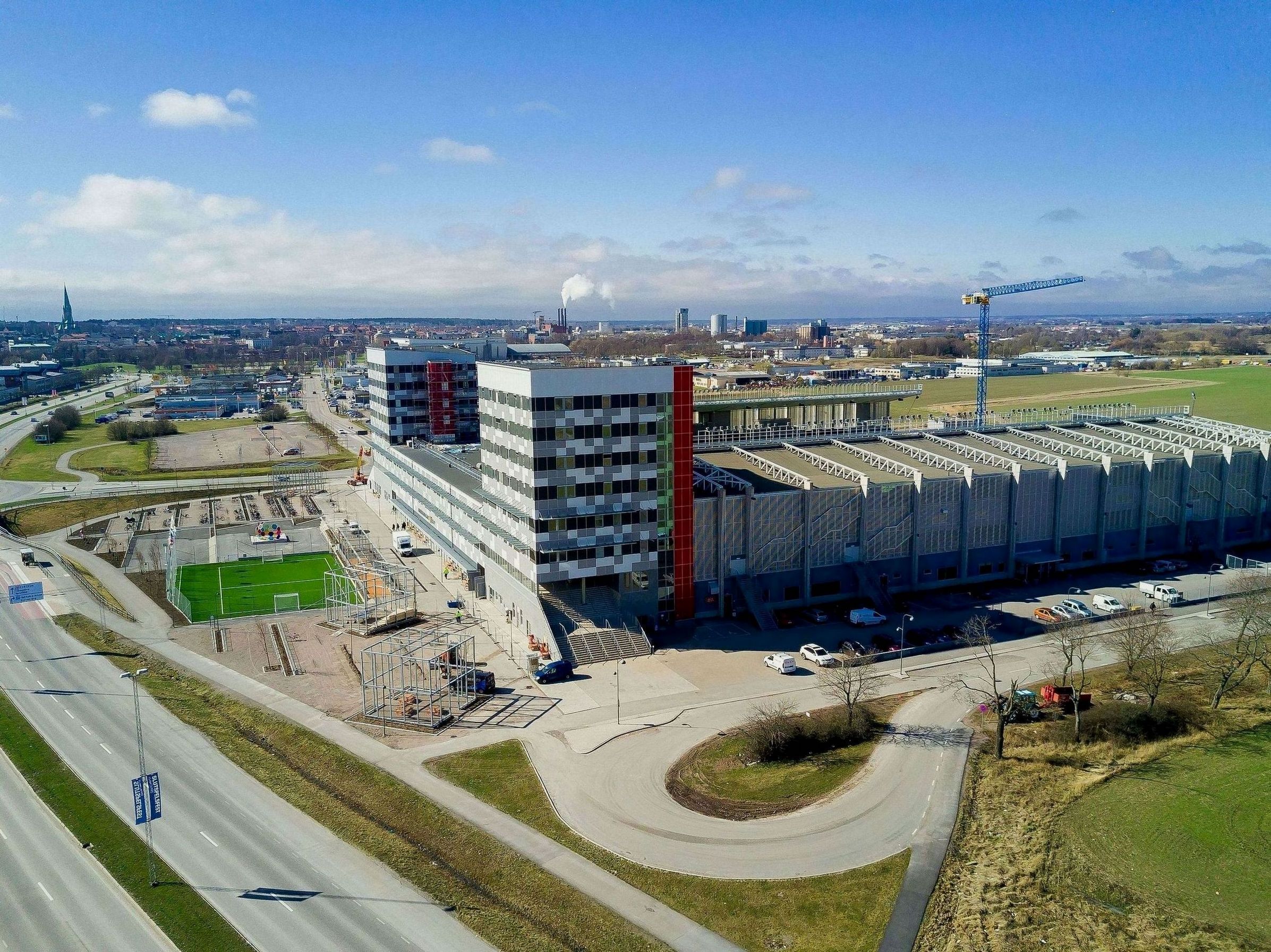 Un grande edificio industriale con diversi piani, un'area di parcheggio e un campo verde nelle vicinanze.