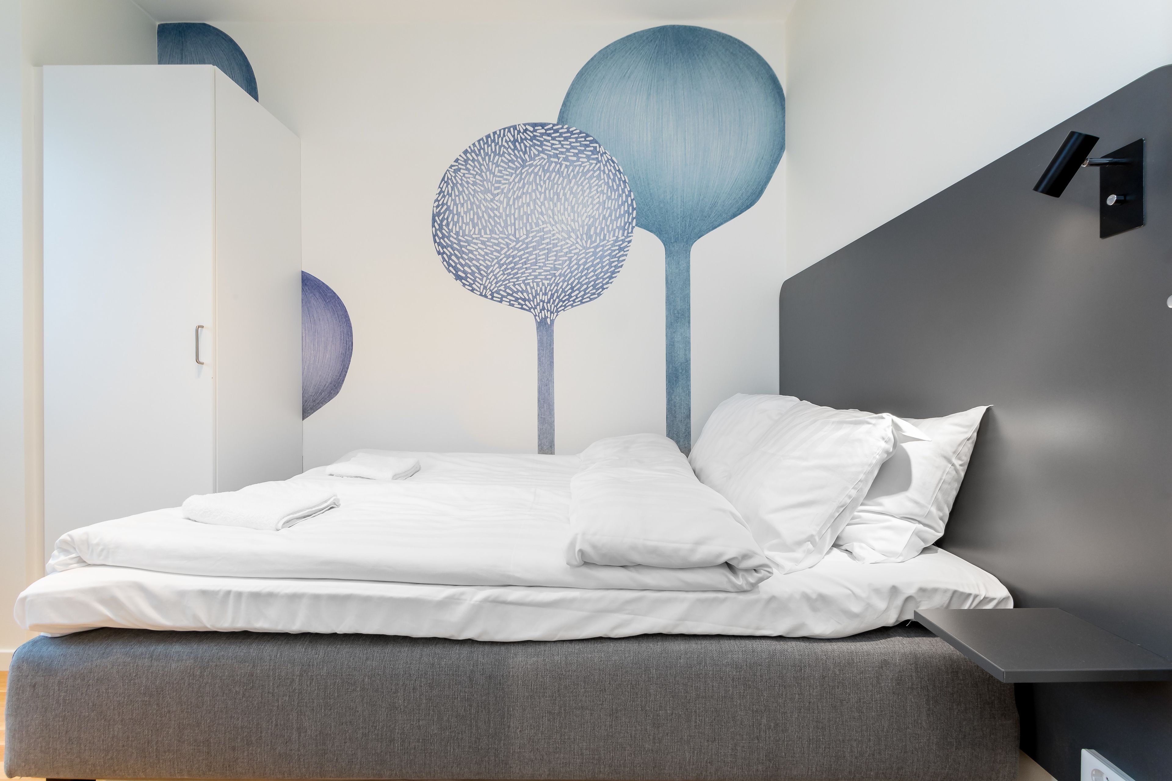 Een netjes opgemaakt bed met witte lakens, een bekleed hoofdbord in grijstint en een wandlamp.