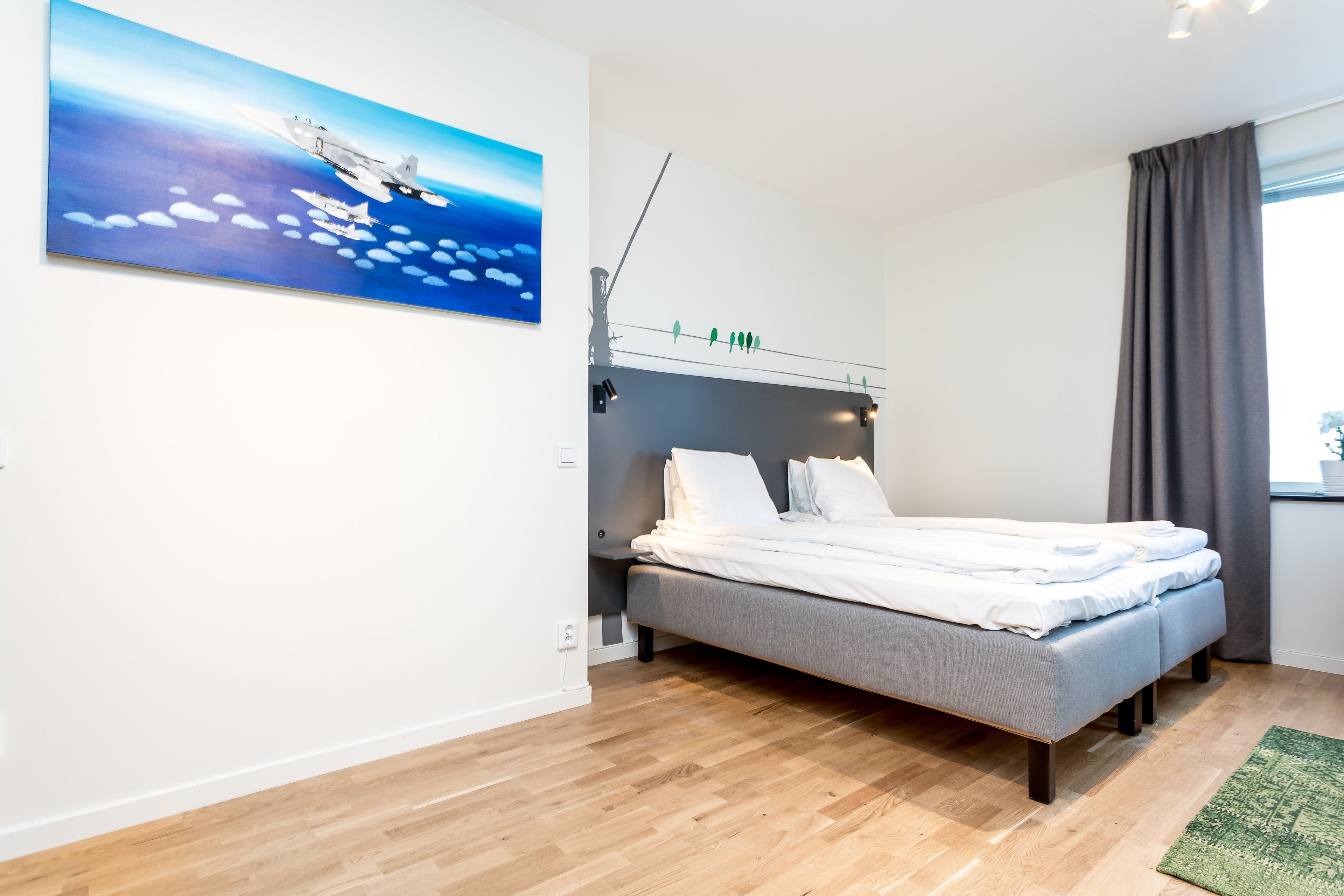 A modern bedroom with a grey bed, white bedding, a painting of an airplane, and a window with curtains.