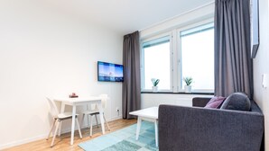 Een moderne woonkamer met een grijze bank, een witte tafel en stoelen. Een flatscreen televisie is aan de muur bevestigd, en er is een raam met gordijnen.