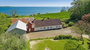 A farmhouse with a large barn and a house with a sloped roof, surrounded by lush greenery and a view of the sea.
