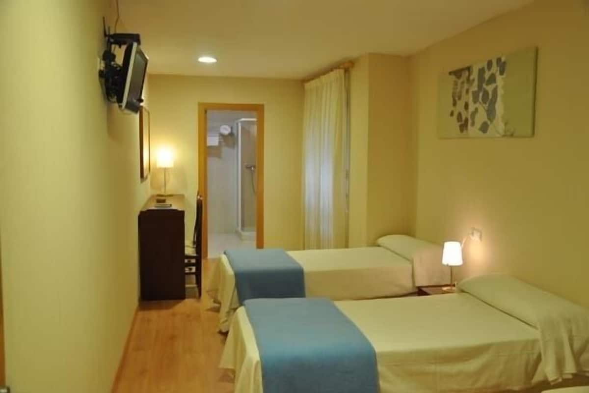 A hotel room with two single beds, a desk with a lamp, a television mounted on the wall, and a shower area visible through a doorway.