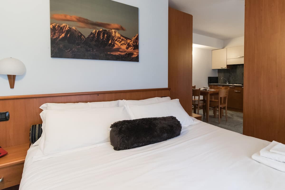 A hotel room with a neatly made bed, a painting of mountains on the wall, a dining area, and a wooden door.