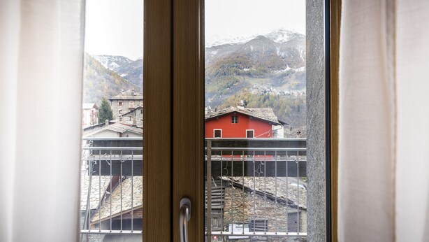 A window with a view of a red building and mountains in the background. - Residence Pizzo Scalino (Chiesa in Valmalenco)