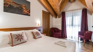 A hotel room with a large bed, two red and white patterned pillows, a wooden headboard, a red armchair, a wooden chair, a painting of mountains, and a window with curtains. - Residence Pizzo Scalino (Chiesa in Valmalenco)