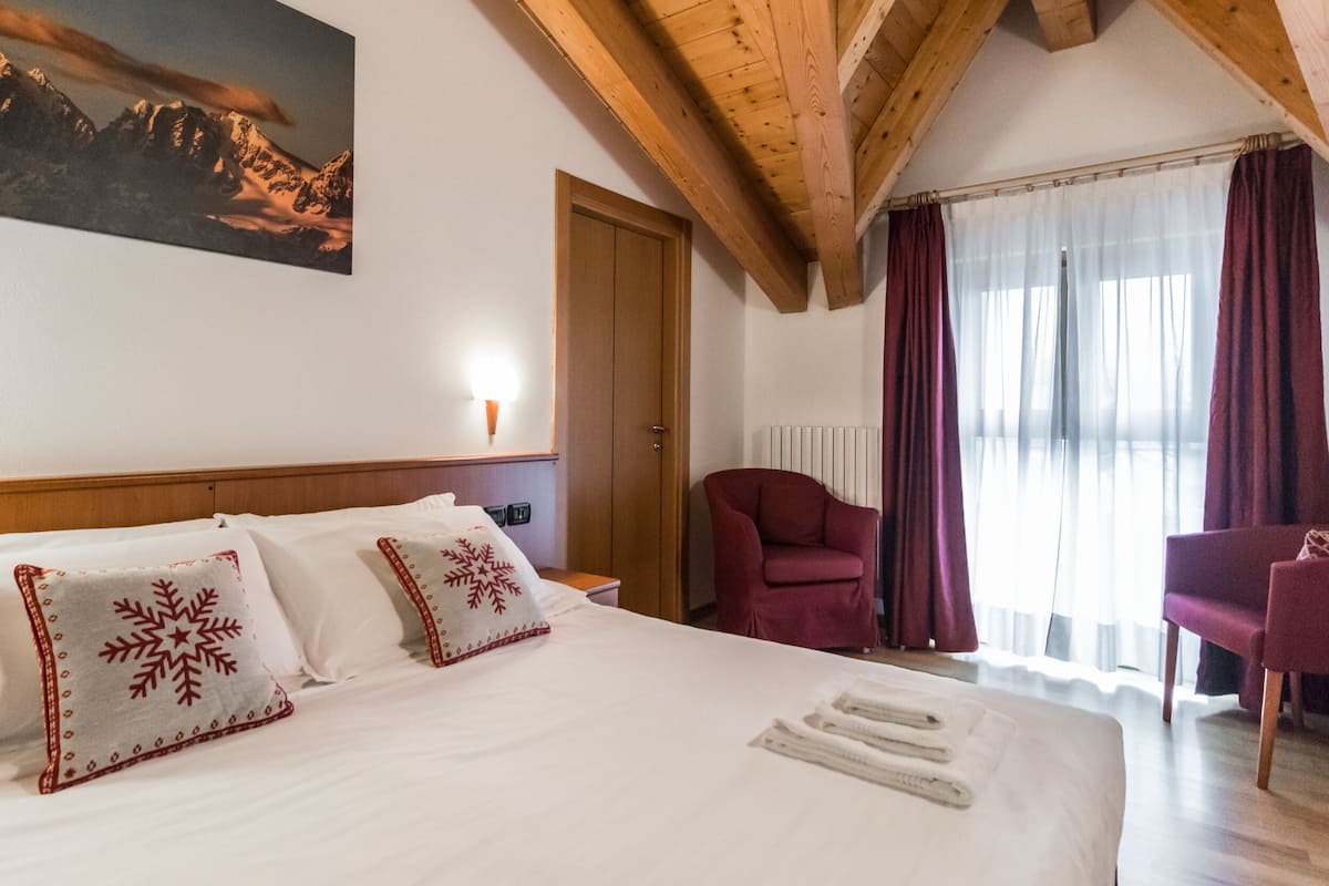 A hotel room with a large bed, two red and white patterned pillows, a wooden headboard, a red armchair, a wooden chair, a painting of mountains, and a window with curtains.