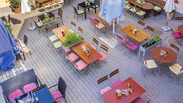 An outdoor café with multiple wooden tables and chairs, some with umbrellas.