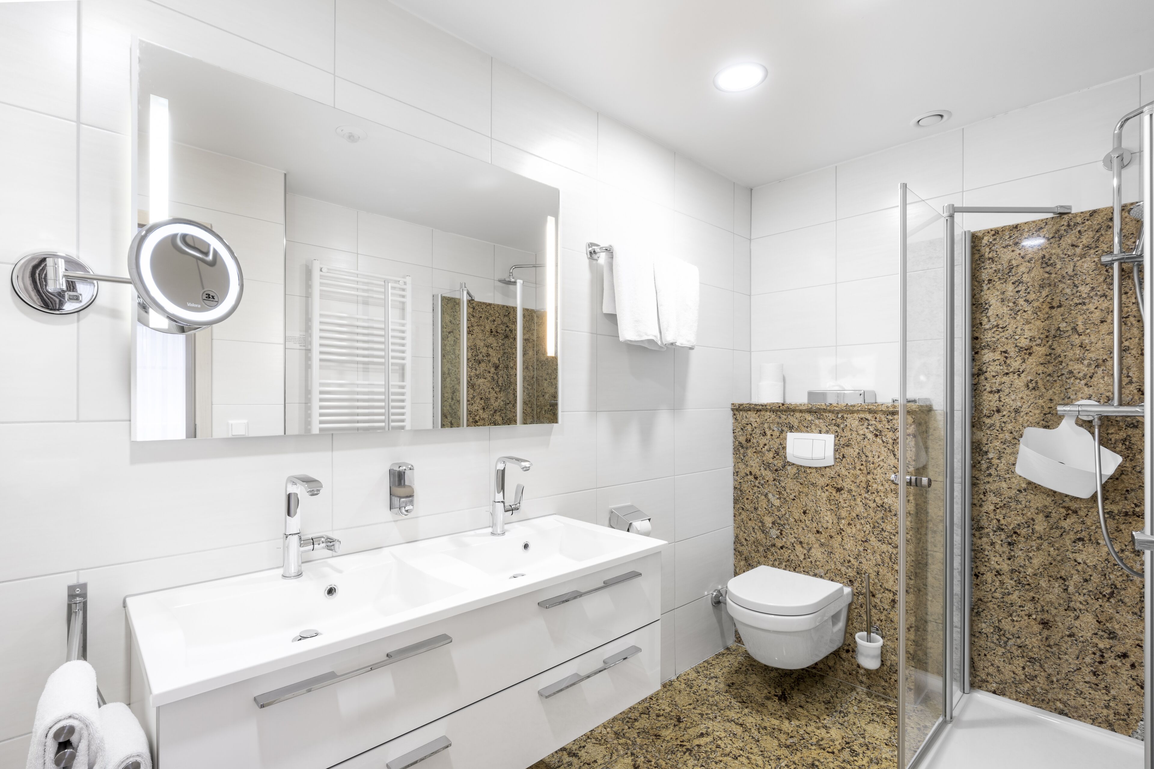 Un baño moderno con tocador blanco, dos lavamanos, un espejo grande, inodoro y cabina de ducha con mampara de vidrio.