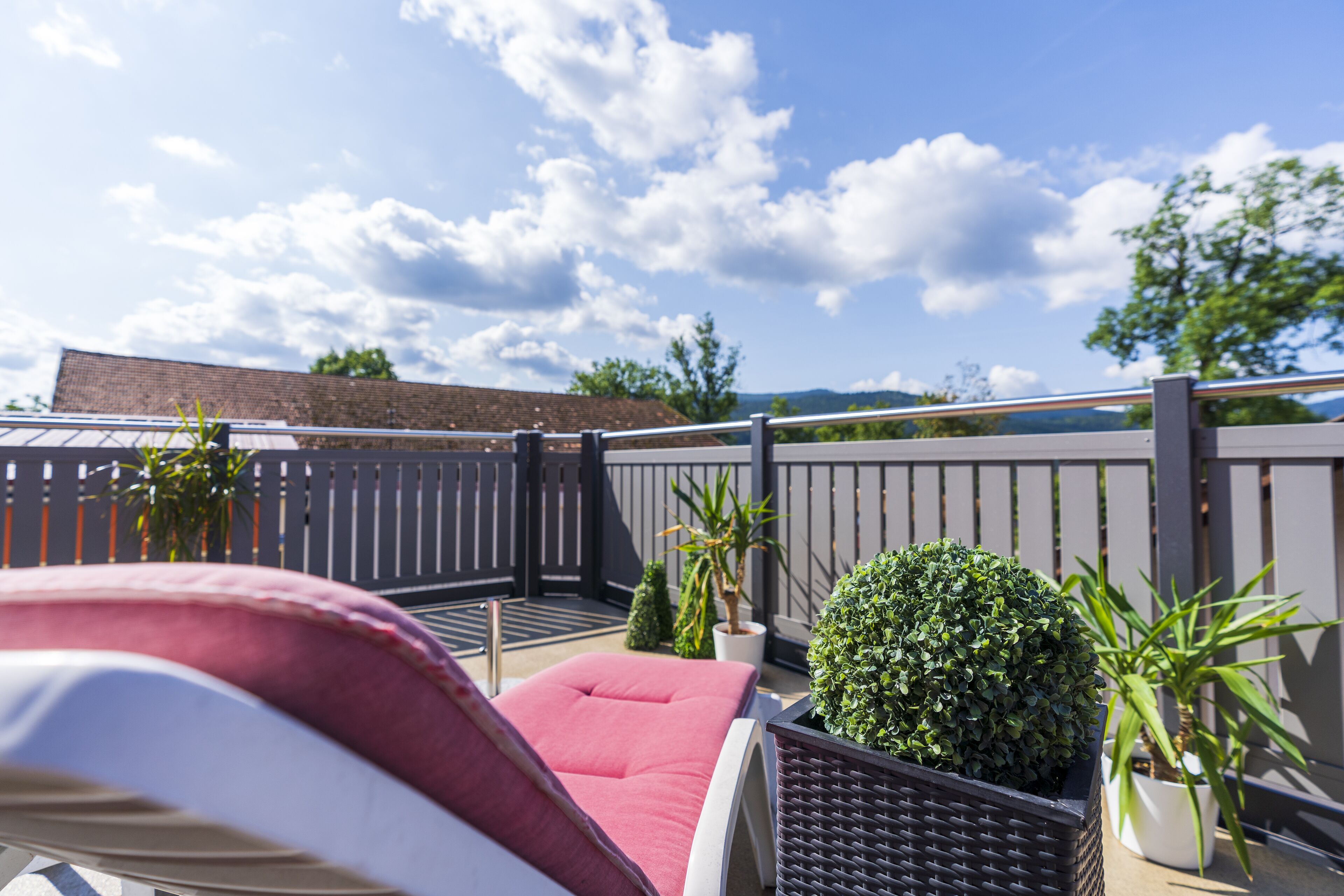A rooftop terrace with a red lounge chair, potted plants, and a view of the sky and distant buildings.