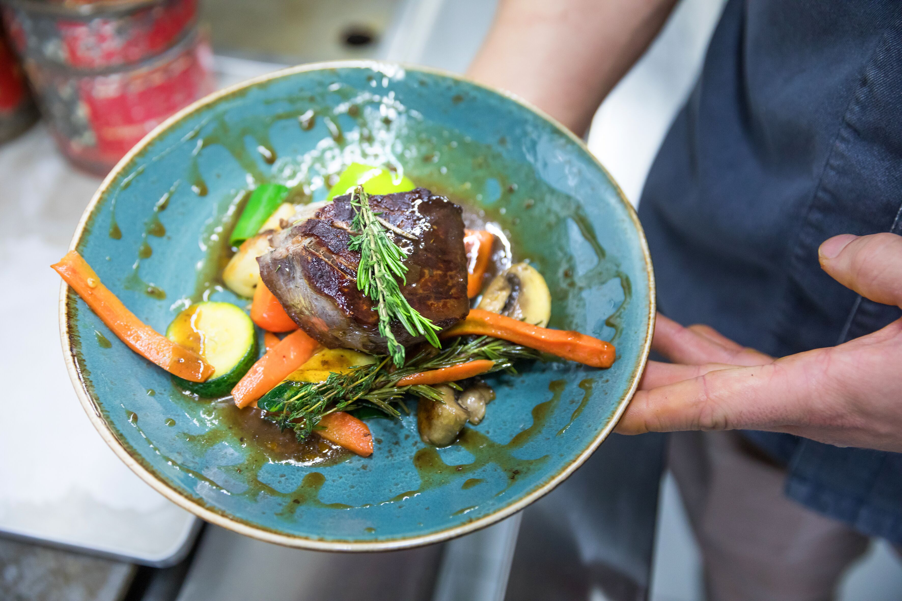 A person holding a plate with a piece of meat, vegetables, and a sprig of herb.