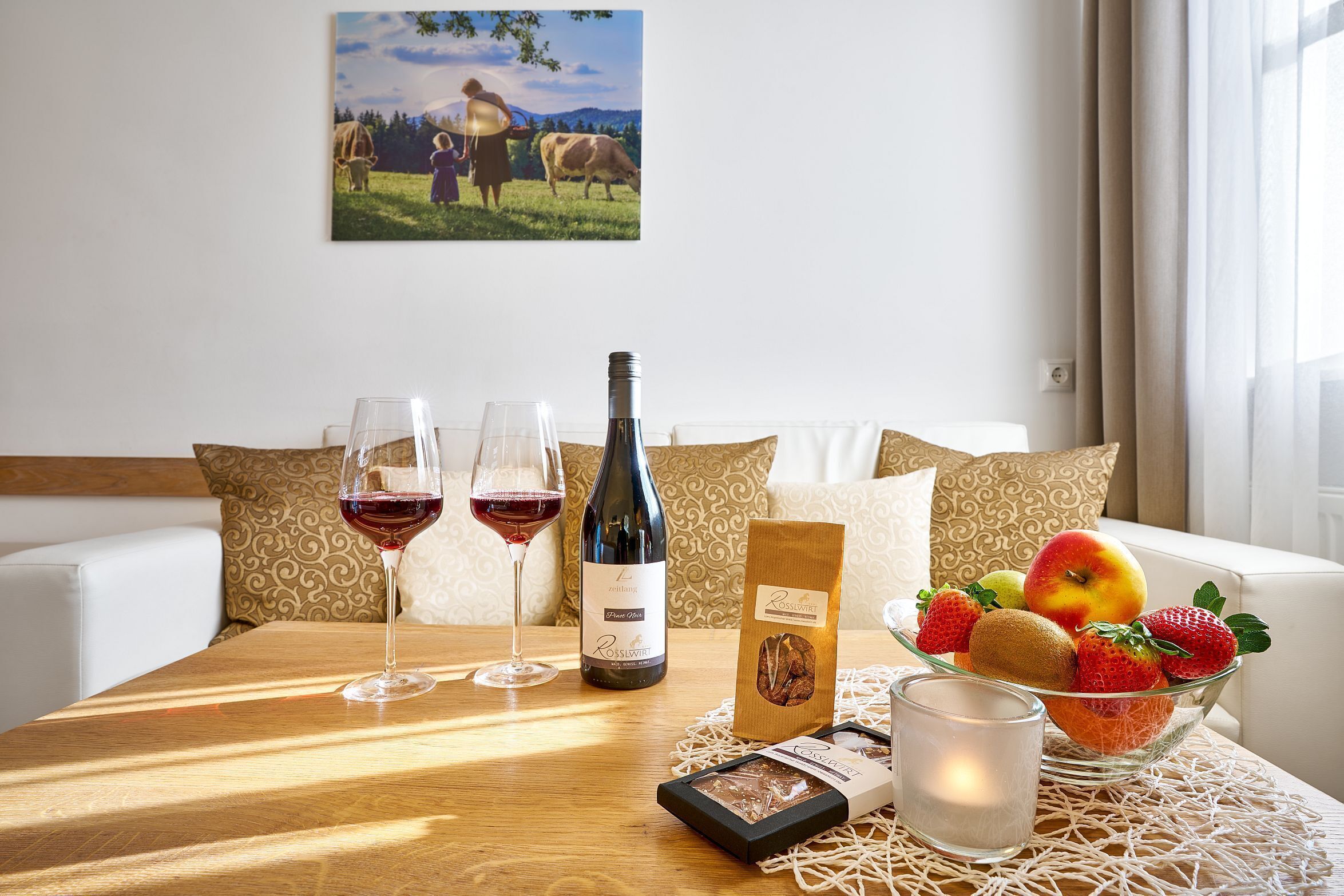 A dining table with a bottle of wine, two glasses, a candle, a fruit bowl, and a box of chocolates.